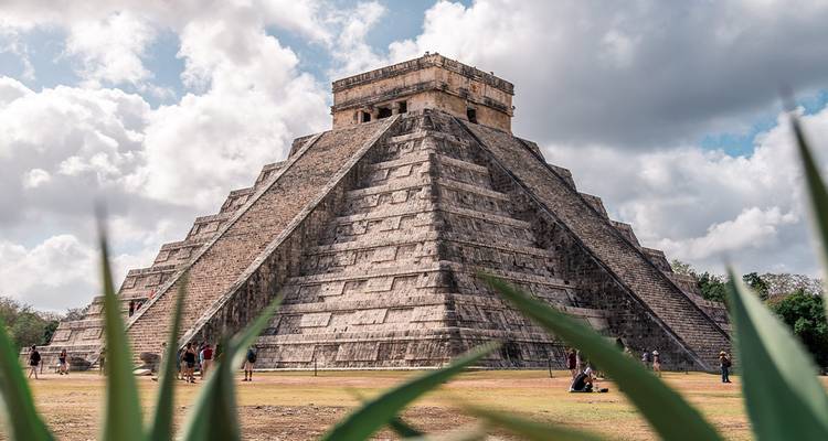 Majestueuse pyramide de Chichén Itzá qui s'élève contre un ciel dramatique encadrée par un feuillage vert.