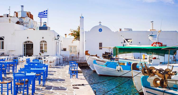 Scène portuaire avec des bâtiments blancs, des chaises bleu vif et des bateaux de pêche arborant le drapeau grec.