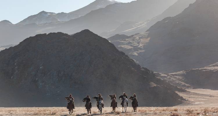 Cazadores de águilas a caballo avanzan por un polvoriento valle bajo montañas dentadas bañadas en una luz suave.