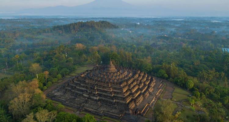 Aerial sunrise view of Borobudur temple emerging from jungle mist with volcano silhouettes beyond.