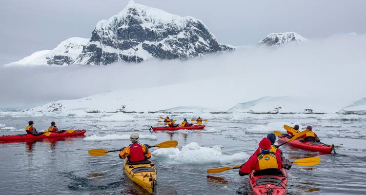 Los kayakistas de colores brillantes reman a través de las aguas heladas de la Antártida con imponentes picos nevados de fondo.
