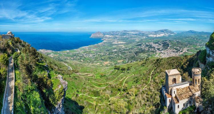 Vista panorámica expansiva desde Erice que muestra valles verdes, costa y montañas distantes bajo un cielo azul despejado.