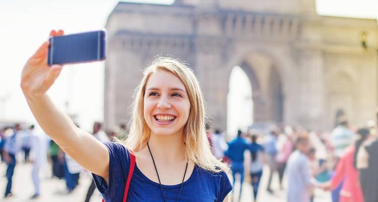 Smiling woman takes a selfie in front of a grand stone arch with bustling crowd behind.
