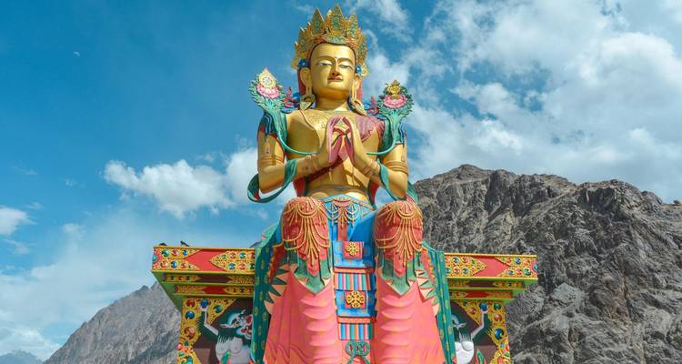 Colorful giant statue of Maitreya Buddha set against rocky Himalayan mountains and blue sky.