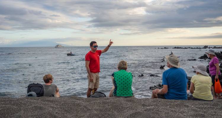 Un guía hace gestos a turistas sentados en arena volcánica con el océano y un yate al fondo.