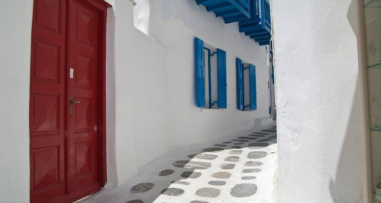 Callejón cicládico blanco encalado y estrecho con una puerta roja brillante, persianas azules y un camino de piedra con patrón bajo la luz clara del día.