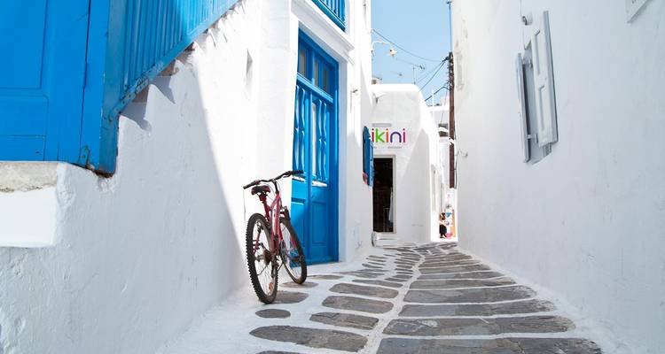 Callejón blanco brillante con puertas azules y una bicicleta rosa apoyada en la pared bajo la luz del día clara.