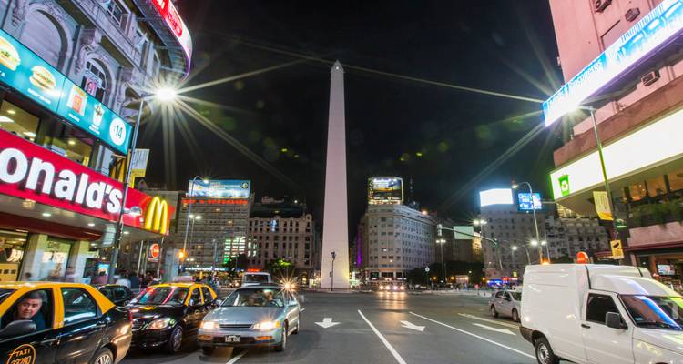 Vista nocturna del Obelisco rodeado de luces brillantes, tráfico y vallas publicitarias en Buenos Aires.