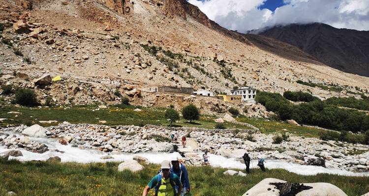 Trekkers wade across stepping stones above a glacial stream below barren Himalayan slopes.