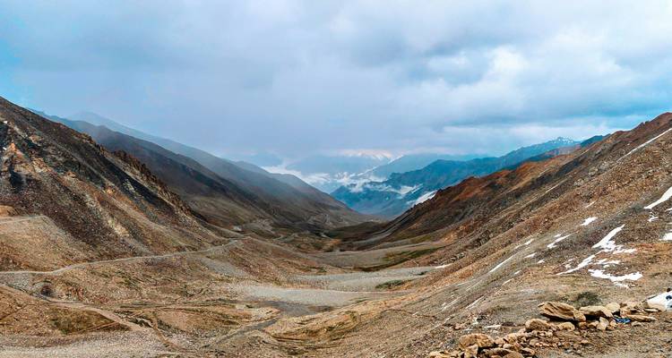 Vaste vallée himalayenne sauvage avec des sentiers sinueux sous des nuages changeants et des pics enneigés au loin.