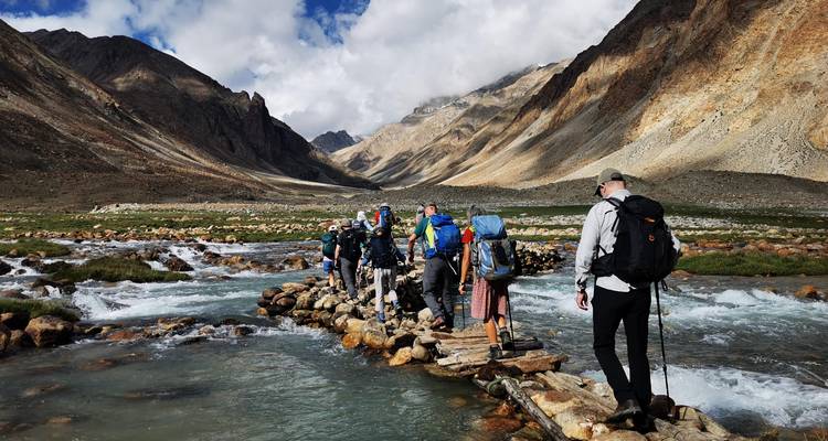 Des randonneurs avec des sacs à dos traversent un pont de pierre étroit au-dessus d'une rivière glaciaire à débit rapide dans une vallée spectaculaire.
