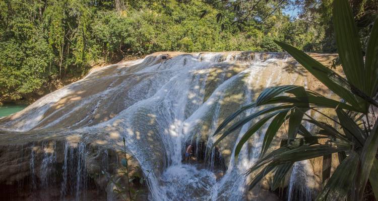 Cascada de múltiples niveles cayendo sobre salientes de piedra caliza suave en la selva espesa, con una persona sola cerca de la base.