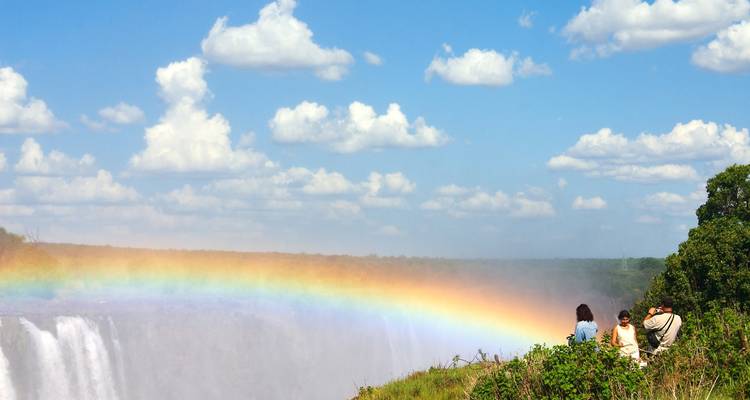 Een levendige regenboog boven een krachtige waterval terwijl drie bezoekers op de rotsrand zitten onder een heldere blauwe lucht met wolkjes.