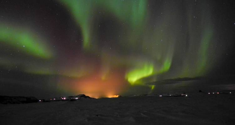 Des aurores boréales vert vif et orange tourbillonnent dans un ciel nocturne arctique étoilé au-dessus d'un sol enneigé.
