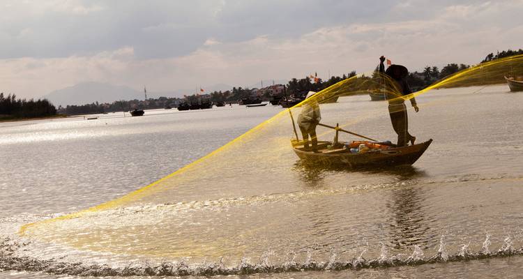 Pescadores lanzando una red dorada desde una pequeña barca de madera en un río muy ancho.