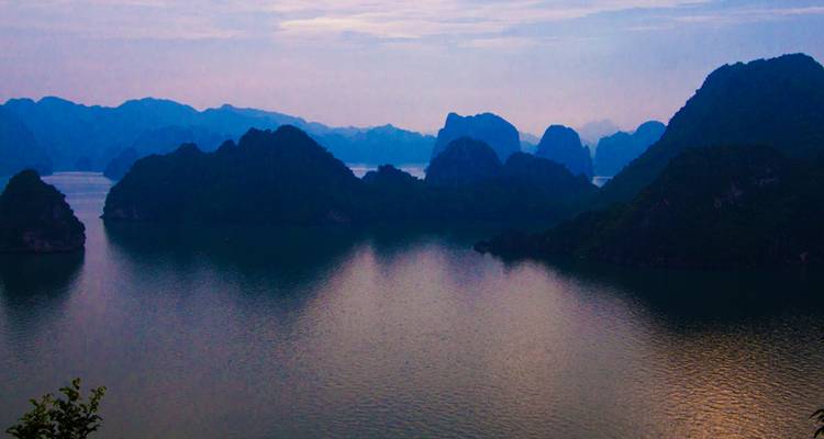 Islas de karst envueltas en niebla elevándose desde aguas tranquilas en la Bahía de Halong al atardecer.