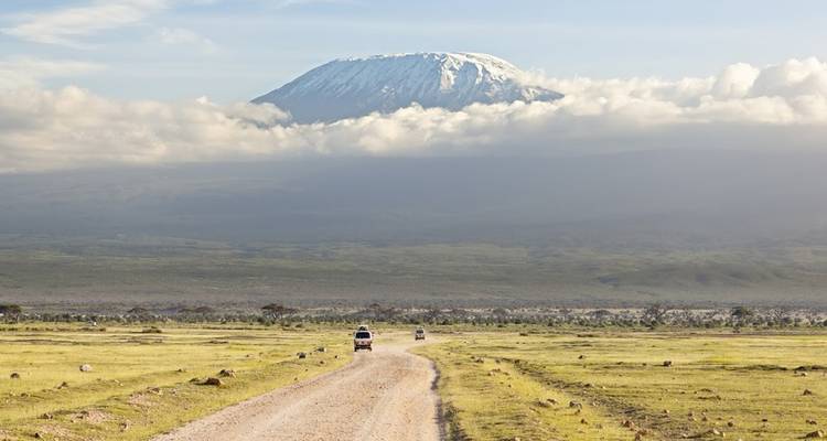 A dirt road leading to Mount Kilimanjaro with clouds and savanna plains.