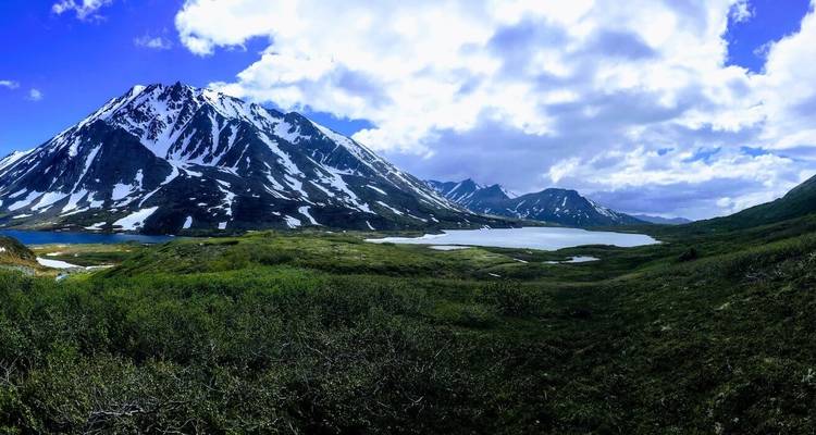 Uitgestrekt landschap van besneeuwde bergen, meren en groen onder een levendige hemel.