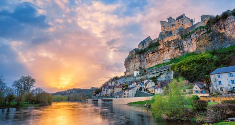 Una puesta de sol ardiente ilumina un castillo medieval en lo alto del acantilado y un pueblo junto al río, reflejando tonos cálidos en el agua tranquila de abajo.
