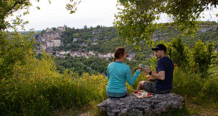 Una pareja disfruta de un picnic con vino y queso en un acantilado rocoso que domina la dramática aldea de montaña de Rocamadour.