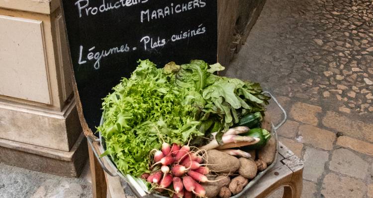 Una canasta de exhibición de mercado rebosa de lechuga fresca, rábanos, papas y calabacín junto a un menú en pizarra en francés.