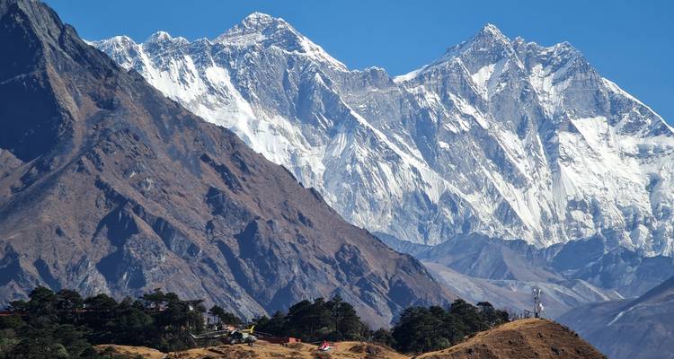 Massive white-clad Himalayan giants, including Mount Everest, dominate the skyline above forested lower ridges under a vivid blue sky.