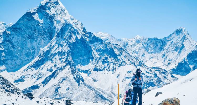Two trekkers with backpacks pause on a snowy slope beneath dramatic Himalayan peaks gleaming in bright sunlight.