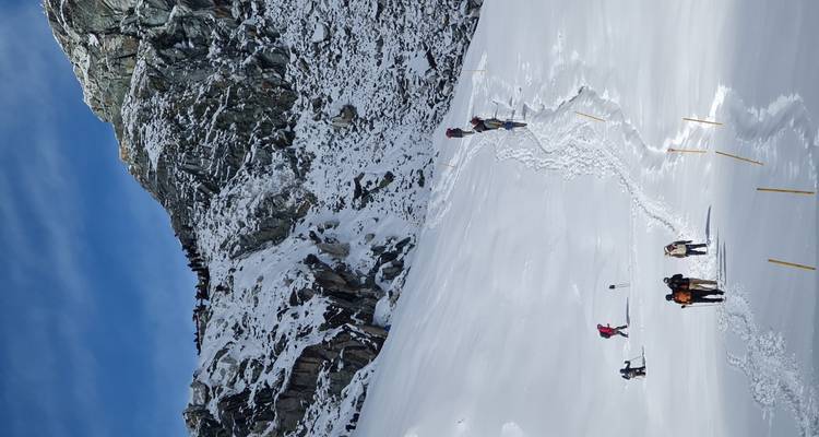A line of climbers trails up a steep, snowy mountainside toward a rocky ridge, leaving deep footprints in the fresh snow.