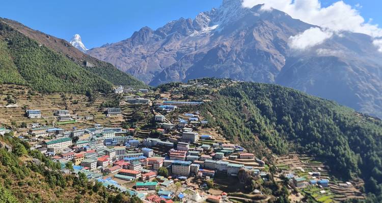 Colourful terraced buildings of Namche Bazaar cling to a green hillside with soaring snow-covered mountains behind.