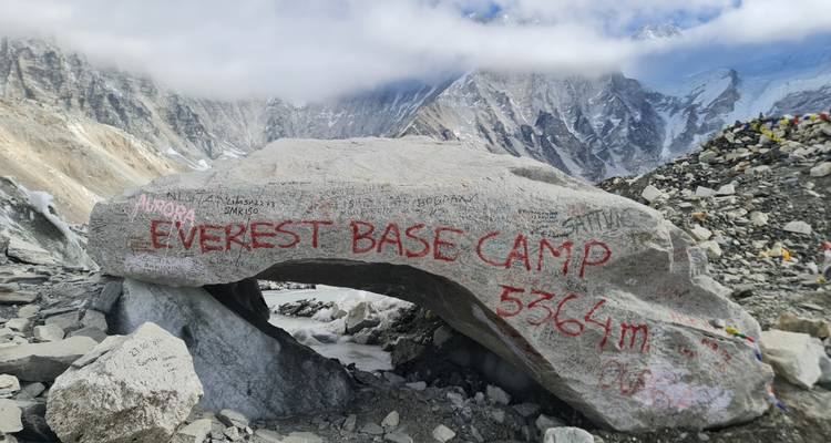 A large boulder at Everest Base Camp painted with red lettering "EVEREST BASE CAMP 5364m" stands among rocky glacial debris.
