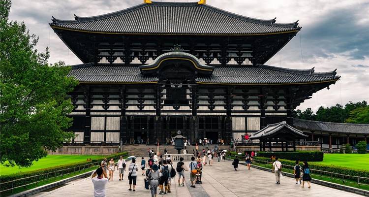 Los visitantes se reúnen frente a la imponente Sala del Gran Buda de madera del templo Todai-ji, con su techo de tejas barrido enmarcado por vegetación.