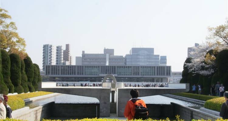 Los turistas observan el cenotafio y el estanque de reflexión del Parque Conmemorativo de la Paz de Hiroshima con el museo moderno al fondo.