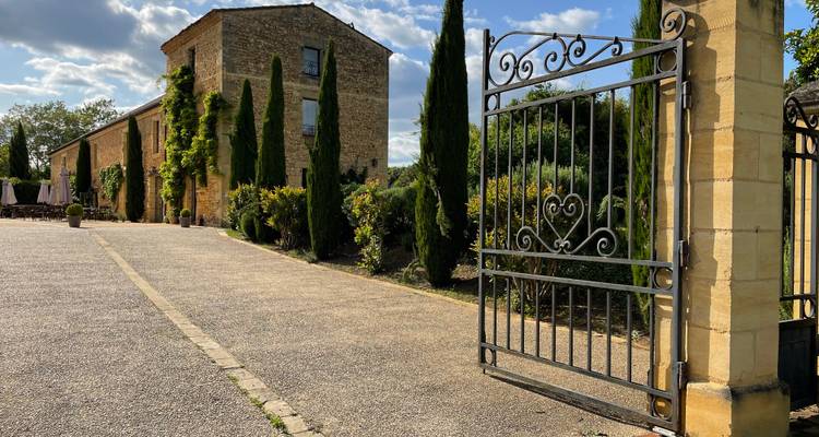 Una puerta de hierro forjado abierta da la bienvenida a los visitantes a una casa de campo de piedra rodeada de altos cipreses.