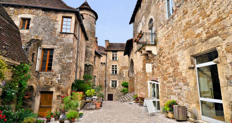 Callejón estrecho de adoquines flanqueado por casas de piedra rústicas con flores y contraventanas de madera en un pueblo francés.