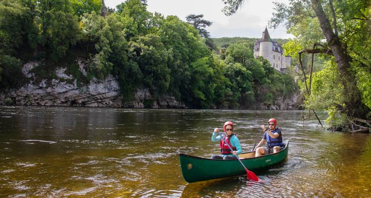 Dos remeros navegan en canoa por un río tranquilo debajo de un acantilado boscoso coronado por un castillo de cuento de hadas.