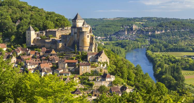 Un château médiéval perché sur une colline surplombe un groupe de maisons en pierre et une rivière sinueuse dans une campagne verdoyante.