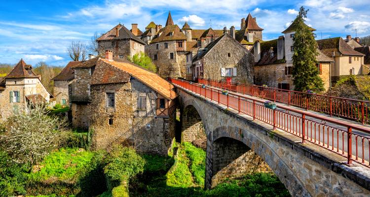 Un pont de pierre aux rambardes rouges mène à un charmant village médiéval construit de maisons en pierre de teinte chaleureuse.