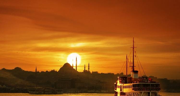Ferry navegando con un atardecer de fondo, con minaretes visibles en la distancia.