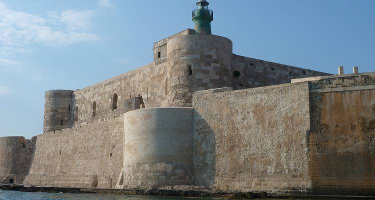 Fortaleza costera de piedra con un faro de techo verde que se eleva sobre murallas desgastadas junto al mar.