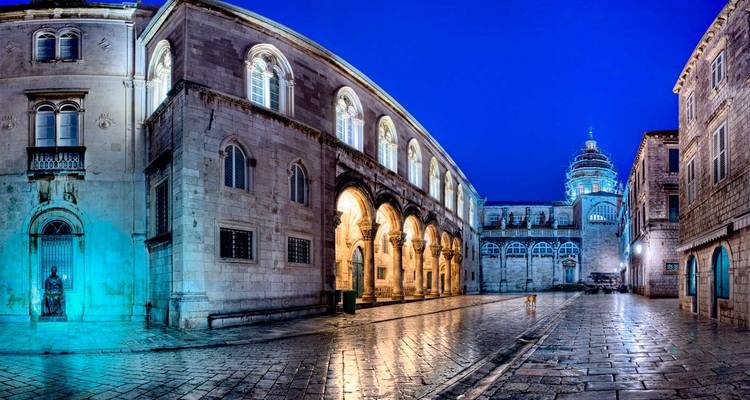 Escena callejera de la hora azul en la ciudad vieja de Dubrovnik con columnatas arqueadas iluminadas por lámparas cálidas sobre adoquines mojados.