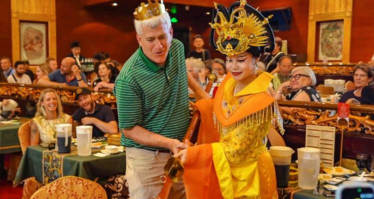 Un comensales que lleva una corona comparte un momento divertido con un artista vestido al estilo de la dinastía Tang durante un espectáculo de banquete.