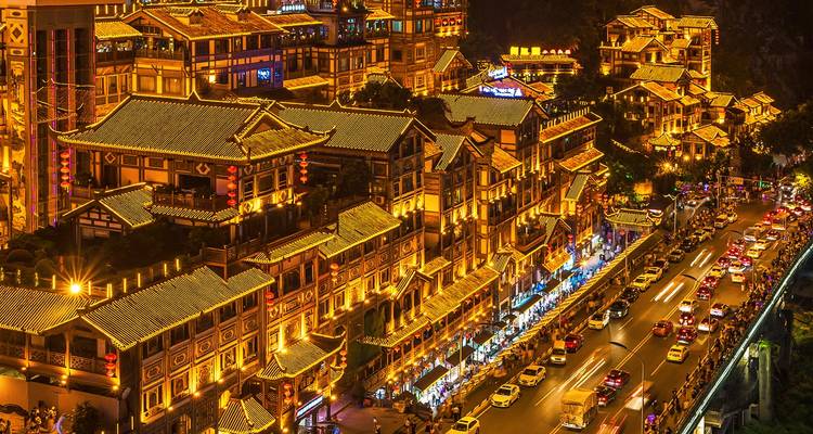 Vue nocturne de bâtiments sur pilotis ornés et brillamment éclairés en cascade le long d'une berge fluviale à côté d'une route animée de Chongqing.
