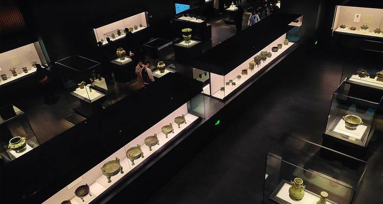 Intérieur d'une galerie de musée moderne sombre avec des vitrines en verre exposant des artefacts anciens et des visiteurs qui se promènent.