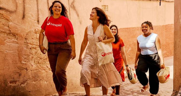 Four women walk laughing through Marrakech’s terracotta alley carrying market totes.