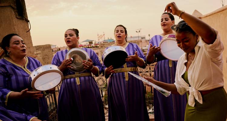 Traditional Moroccan female musicians in violet dresses drum and sing on a rooftop at dusk.