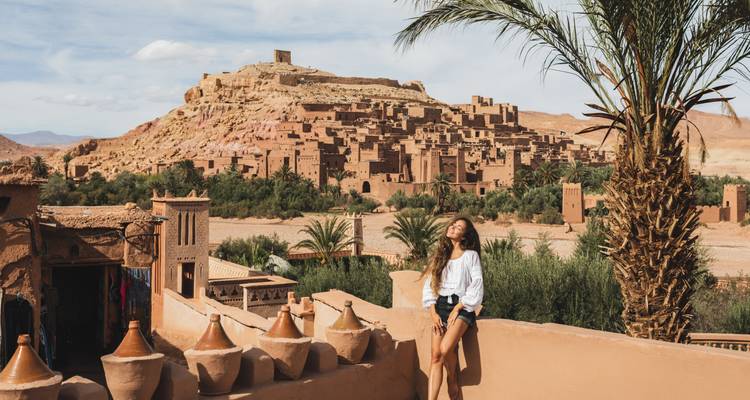 Young female traveler in white blouse and shorts relaxing on terrace overlooking the mud-brick ksar of Ait Benhaddou.