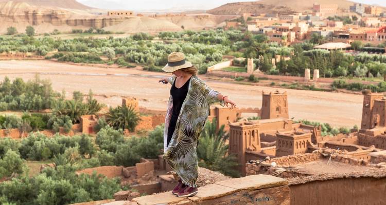 Woman balancing on ancient adobe wall with lush oasis and earthen village of Ait Benhaddou behind her.