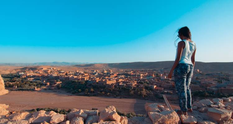 Girl standing on rocky ridge gazing over vast arid valley dotted with clay houses under bright blue sky.