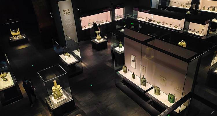 Halfdonker museumzaal met oude jade, brons en aardewerk artefacten in stijlvolle glazen vitrines.
