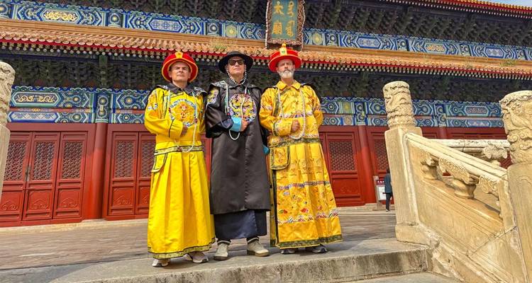 Tres visitantes vestidos con trajes imperiales brillantes posan con orgullo frente a las puertas del palacio rojo adornado en Pekín.